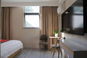 Ett hotellrum med en säng, ett skrivbord, en stol, ett litet bord med en växt och ett fönster med gardiner.