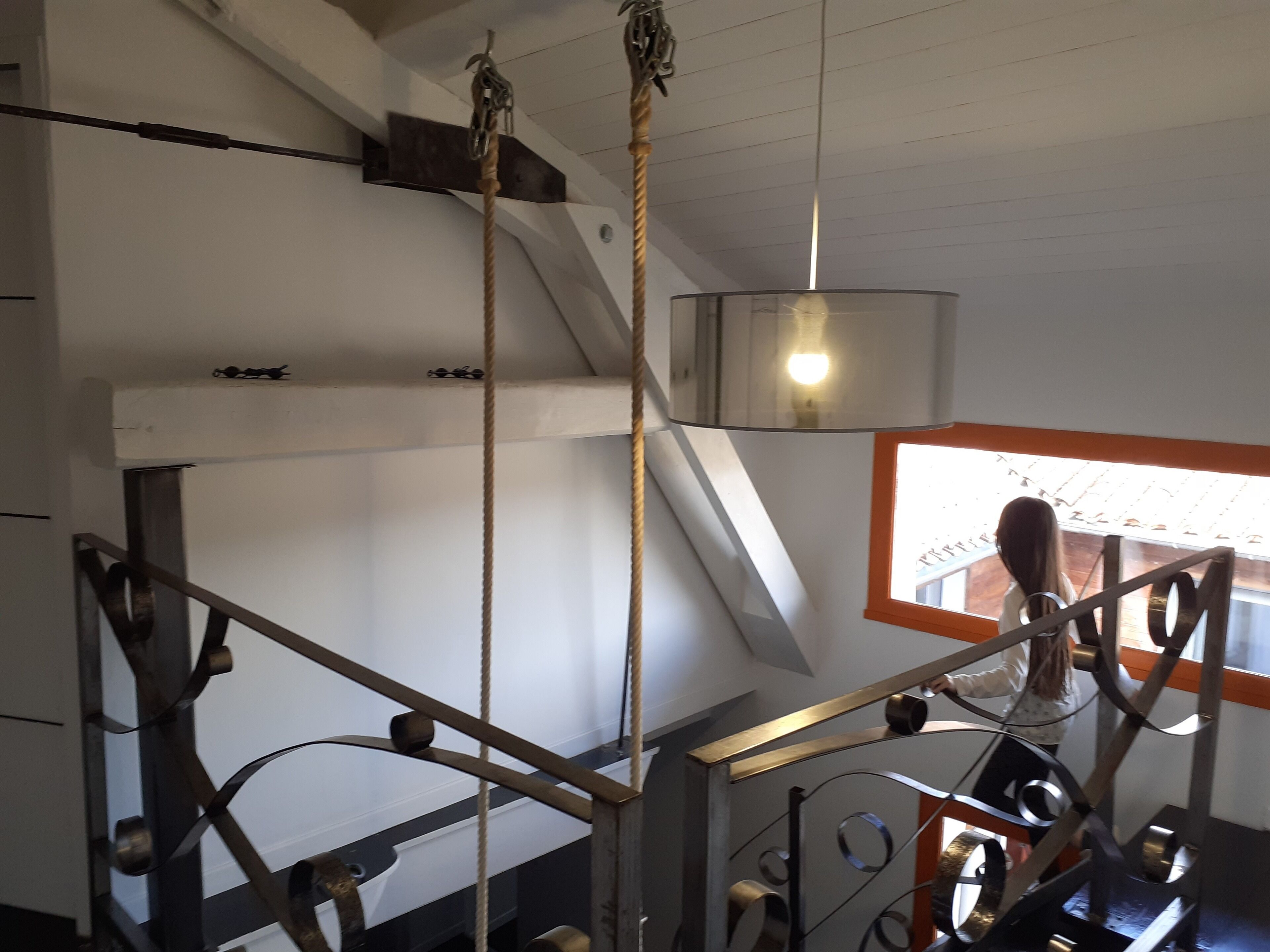 Un escalier avec des rampes en métal et une corde servant de support, une fenêtre avec un cadre orange et un luminaire suspendu.