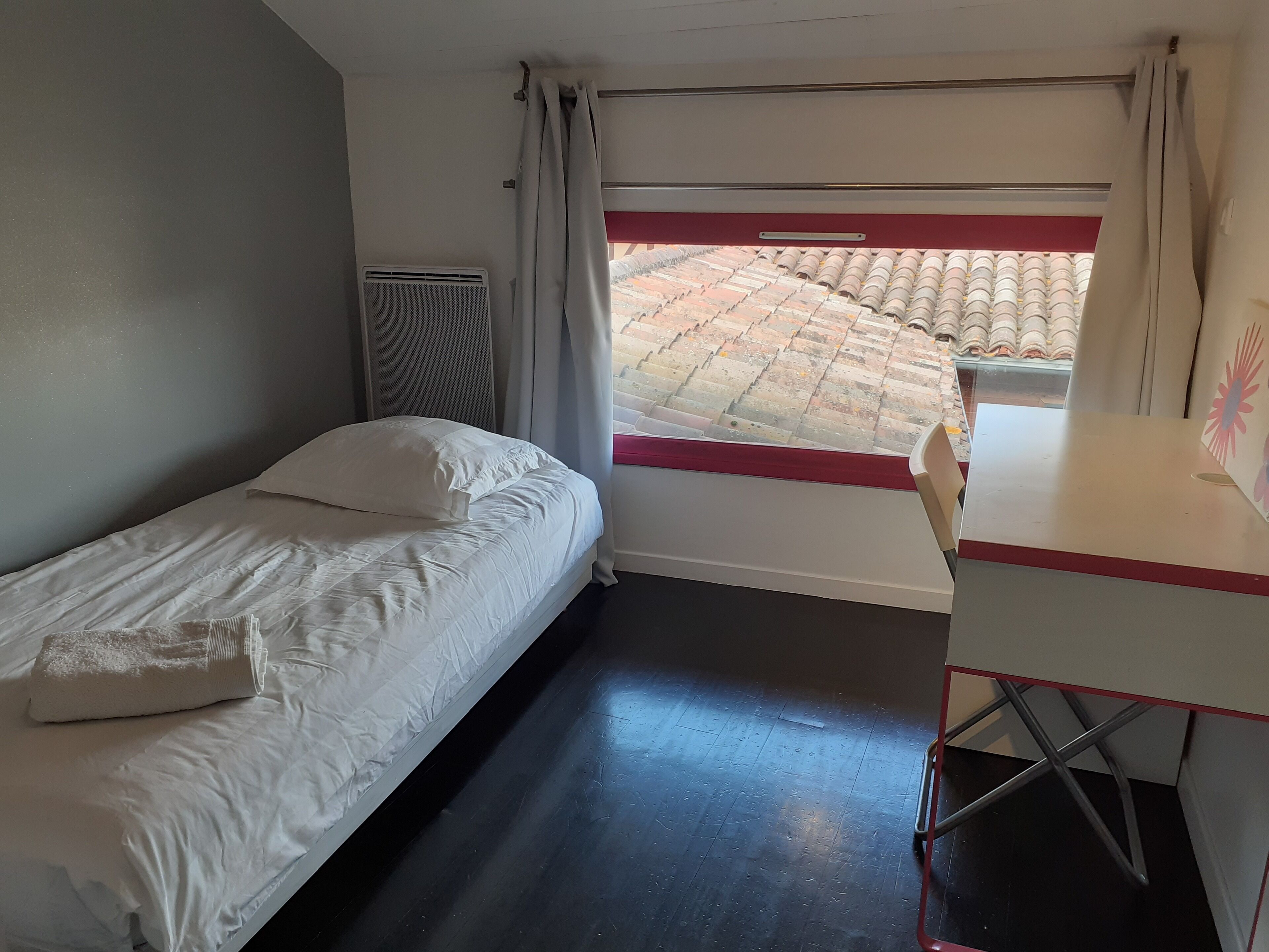 Una habitación pequeña y funcional con una cama individual, un escritorio y una ventana con vistas a un tejado de baldosas.