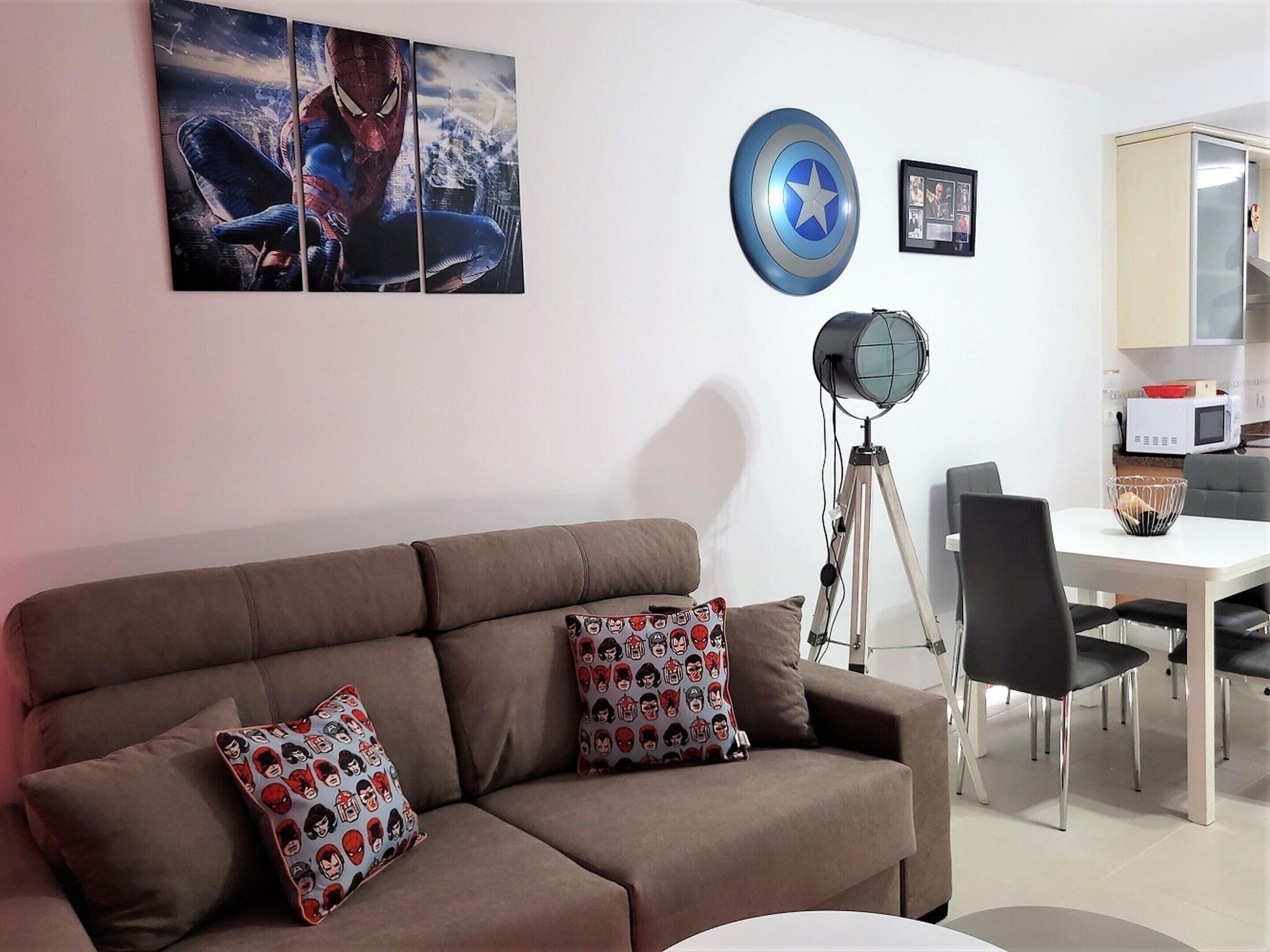 A living room with a brown sofa, two patterned pillows, a large Spider-Man artwork, a Captain America shield, and a floor lamp.