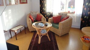 A cozy living room with wicker furniture, a glass coffee table, and a view of a house through a window with floral curtains. - Time out Zu2! Modern Retreat (Cuxhaven)