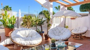A patio area with white cushioned chairs, a glass table, and potted plants.