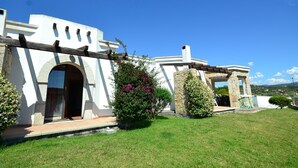 A white building with a stone archway entrance, a pergola, and a small garden area with a bush and flowers. - Villa in Alghero With Pool & Near Town (Alghero)
