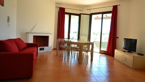 A living room with a red sofa, a fireplace, a dining table with chairs, a TV on a cabinet, and a sliding glass door leading to an outdoor area. - Villa in Alghero With Pool & Near Town (Alghero)