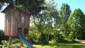 Une cabane en bois avec un toboggan, entourée d’une végétation luxuriante et d’arbres.