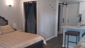 A bedroom with a bed, a bedside table, a nightstand, a window with curtains, and a door leading to a bathroom. - Quaint studio (Largo)