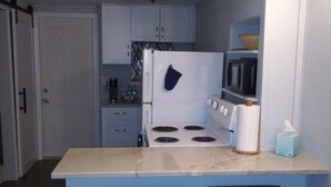 A compact kitchen with white cabinets, a stove, a microwave, and a small bar stool. - Quaint studio (Largo)