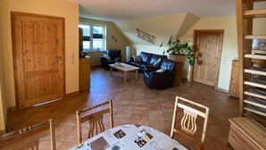 A spacious living area with a wooden floor, a black leather sofa, a coffee table, and a dining table with chairs. There is a large window providing natural light and a door leading to another room. - Inviting Apartment in Pruchten With Garden (Pruchten)