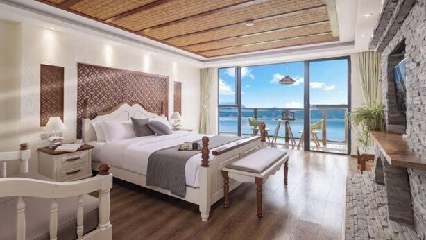 A bedroom with a large bed, a bench, a desk, and a view of the sea.