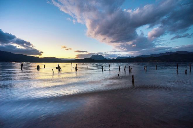 Un lago tranquilo con pilotes de madera en el agua, rodeado de montañas y un cielo al atardecer.