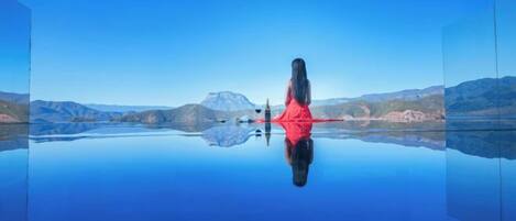 A person in a red top sitting by a calm lake with mountains in the background.