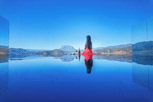 Uma pessoa vestida de vermelho sentada à beira de um lago calmo, com montanhas ao fundo.