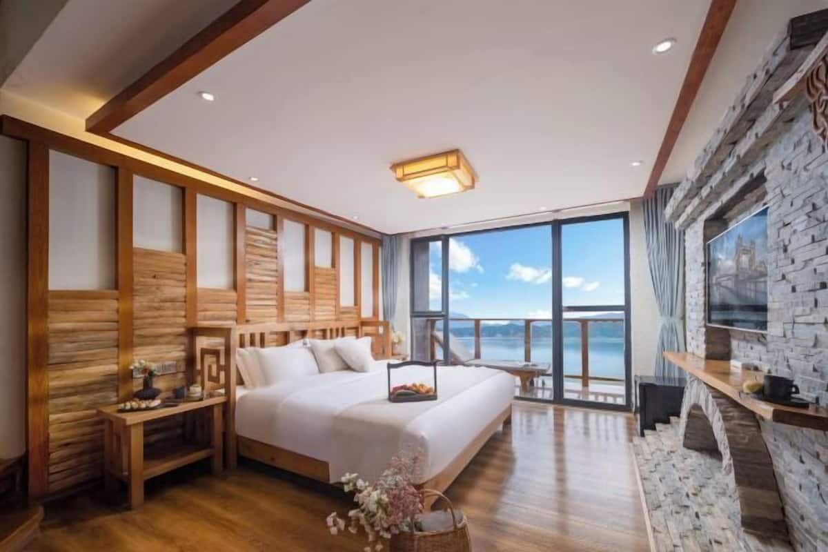 Quarto com uma cama grande, cabeceira de madeira, uma mesa de cabeceira, uma pequena mesa com um vaso e vista para o mar através de janelas amplas.