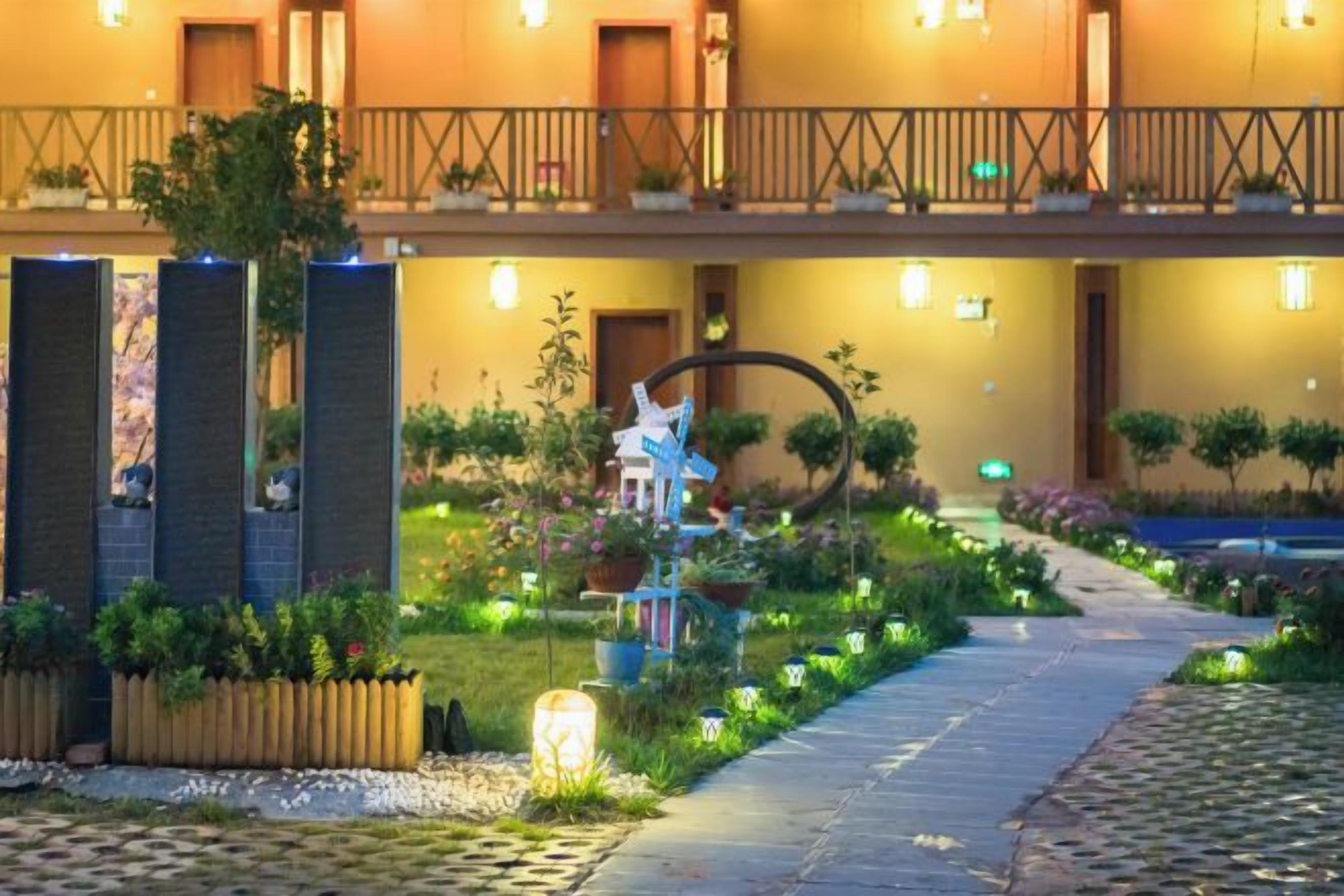 Entrada de un hotel con un sendero que conduce al edificio principal, un pequeño jardín con plantas y una estructura decorativa, y un balcón con barandillas.