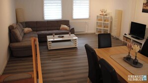 A modern living room with a sofa, coffee table, and dining area. There are two windows with blinds, a TV, and a bookshelf. - Ferienhaus Fuldablick Das Ferienhaus Inmitten der Altstadt von Rotenburg /fulda (Rotenburg an der Fulda)
