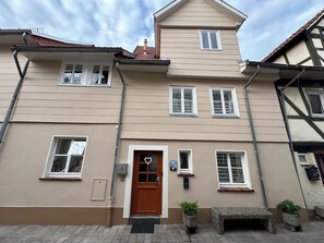 A two-story building with a brown door, multiple windows, and a small porch area with a bench and potted plants.
