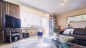 A modern living room with a flat-screen TV, a glass coffee table, a sofa, and a dining area with a table and chairs.