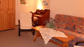 Un salon chaleureux avec un meuble en bois, un fauteuil blanc, un canapé à motifs et une petite table recouverte d’une nappe au crochet.