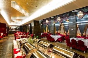 Un salón de banquetes con mesas dispuestas para comer, sillas rojas y una larga barra con una parrilla.