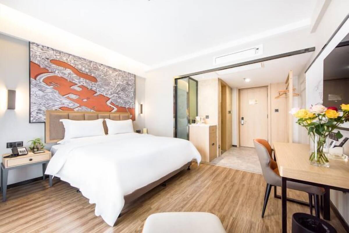 A modern hotel room with a large bed, a dining table with chairs, a small desk, and a large abstract painting on the wall.