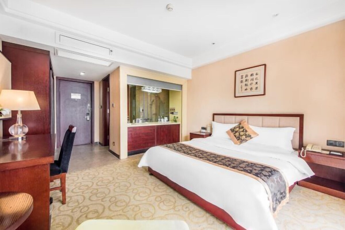 A hotel room with a large bed, a desk, a chair, a mirror, and a bathroom visible through an open door.