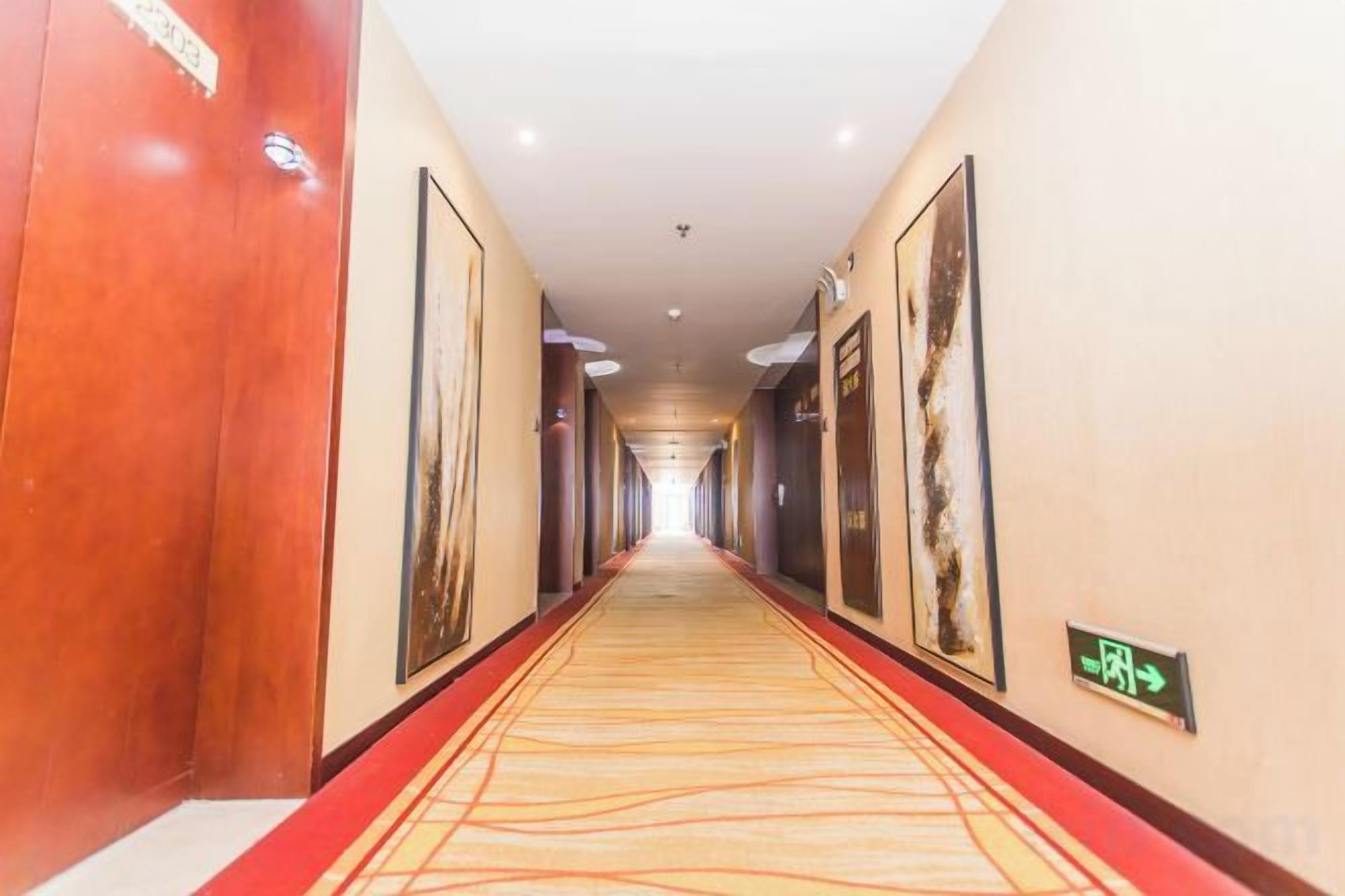 A hotel corridor with a red carpet, framed artwork on the walls, and a directional sign.