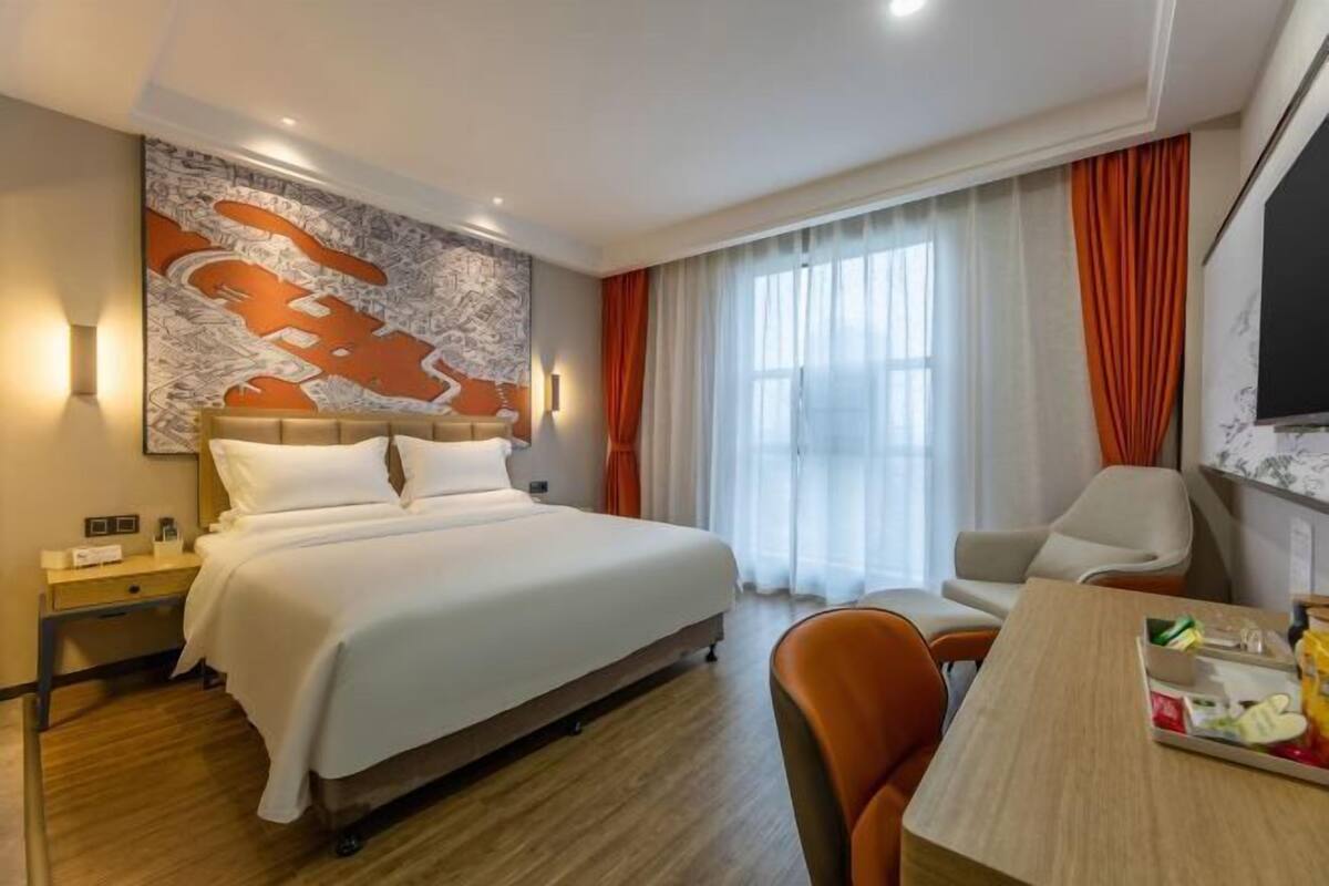 A hotel room with a large bed, a desk with a chair, a television, and a large window with curtains.