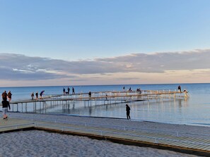 Una playa con un muelle de madera que se adentra en el agua, gente caminando por él y una zona de arena.