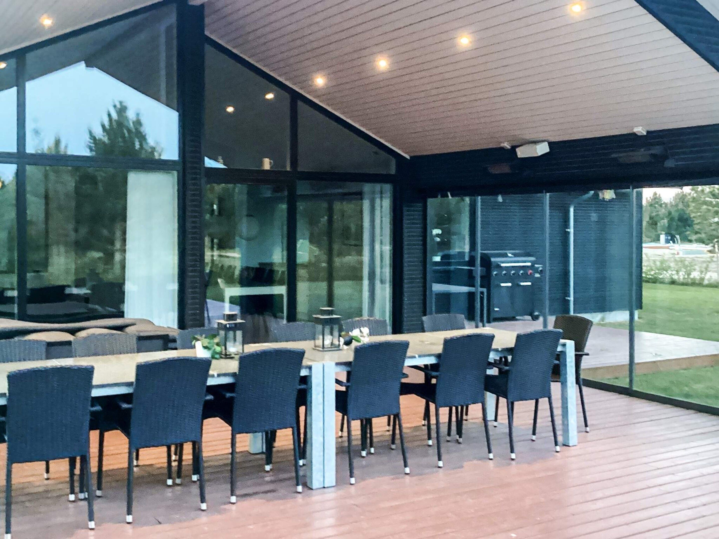 Un área de comedor exterior moderna con terraza de madera, paredes de vidrio y vistas al exterior.