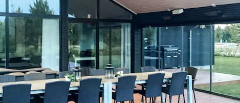 A modern outdoor dining area with a wooden deck, glass walls, and a view of the outdoors.