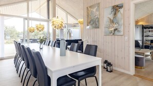 A modern dining area with a long white table, black chairs, and yellow pendant lights. There are large windows providing natural light and a view of the outdoors.