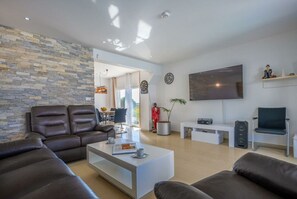 A modern living room with a stone wall, leather sofas, a flat-screen TV, and a dining area visible through an open door.
