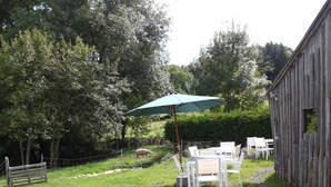 Un área de estar rústica al aire libre con una mesa y sillas bajo un paraguas verde, rodeada por una valla de madera y árboles.
