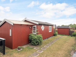 A red wooden building with a small window and a door, a paved pathway, and a grassy yard.