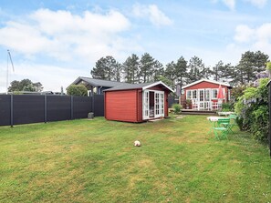 A red wooden shed in a backyard with a lawn, a white fence, and a red house with a porch. - 3 Person Holiday Home in Karrebaeksminde (Karrebæksminde)