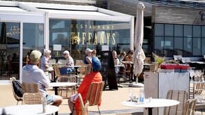 An outdoor seating area of a restaurant with wicker chairs and tables, a covered section with a glass canopy, and a building with large windows in the background. - Apartment in Nieuwpoort by the Beach (Nieuwpoort)