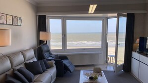 A hotel room with a leather sofa, a coffee table with a fruit bowl, a TV, and a view of the beach. - Apartment in Nieuwpoort by the Beach (Nieuwpoort)