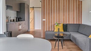 A modern living room with a grey sofa, a small round table, and a kitchen area with grey cabinets and a wooden door. - Chalet in Dutch Park With Garden and Terrace (Hoogersmilde)
