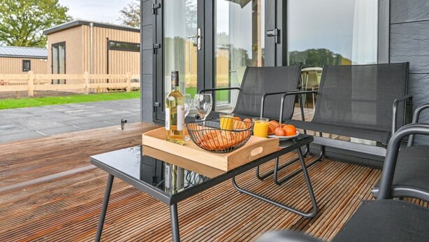 A modern outdoor seating area with a table set for two, featuring a bottle of wine, glasses, a basket of bread, and a plate of oranges. - Chalet in Dutch Park With Garden and Terrace (Hoogersmilde)