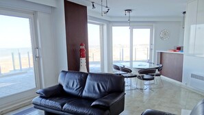 A modern living room with a black leather sofa, a dining area with a table and chairs, and large windows offering a view of the beach. - Seaside Serenity Apartment (Nieuwpoort)