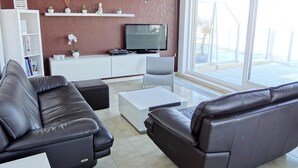 A modern living room with a black leather sofa, a white coffee table, and a flat-screen TV on a white cabinet. There are shelves with decorative items and a large window providing a view outside. - Seaside Serenity Apartment (Nieuwpoort)