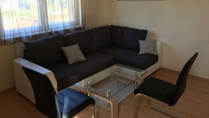 A modern living room with a dark brown sectional sofa, a glass coffee table, two black chairs, and a large window with sheer curtains. - Bolliger 2 Modern Retreat (Kalkhorst)