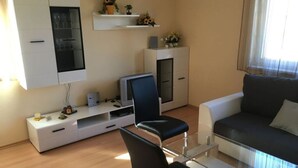 A modern living room with a glass coffee table, a black leather chair, a white sofa, a TV on a white stand, and a small shelf with plants. - Bolliger 2 Modern Retreat (Kalkhorst)