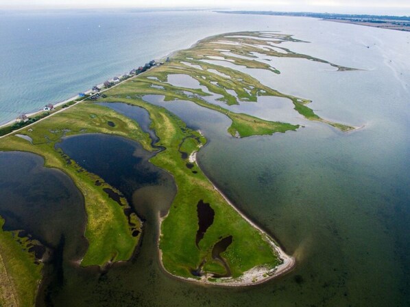 An aerial view of a coastal area with a long, narrow strip of land with a road and buildings along one side, and a series of water bodies and green areas on the other.