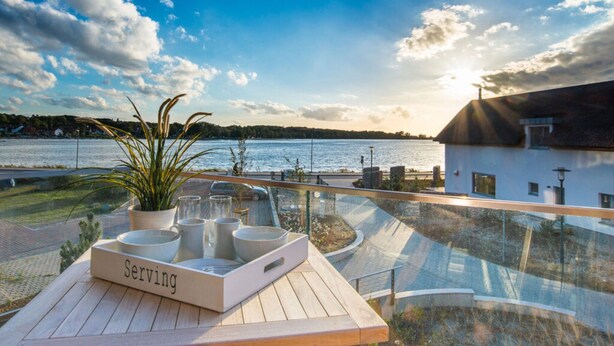 A terrace with a serving tray, potted plants, and a view of a lake and buildings. - Holiday Apartment 11deg East (Heiligenhafen)