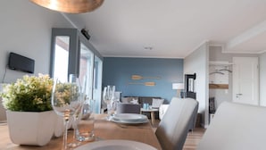 A modern dining area with a wooden table set for two, a blue accent wall, and a view through an open door. - Holiday Apartment 11deg East (Heiligenhafen)
