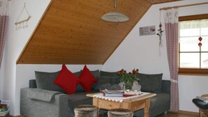 Ein gemütlicher Raum mit schrägverlegtem Holzboden, einem grauen Sofa mit roten Kissen, einem kleinen Holztisch mit einer Vase voller roter Blumen und einem Fenster mit karierten Vorhängen.