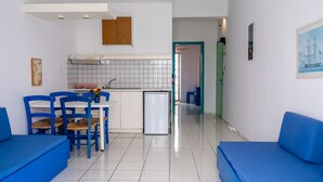 A modern kitchen with white cabinets, a built-in oven, and a sink. There are blue chairs and a dining table with a vase of flowers. A small refrigerator is visible, and the floor is tiled. - Enplo Apartments (Hersonissos)