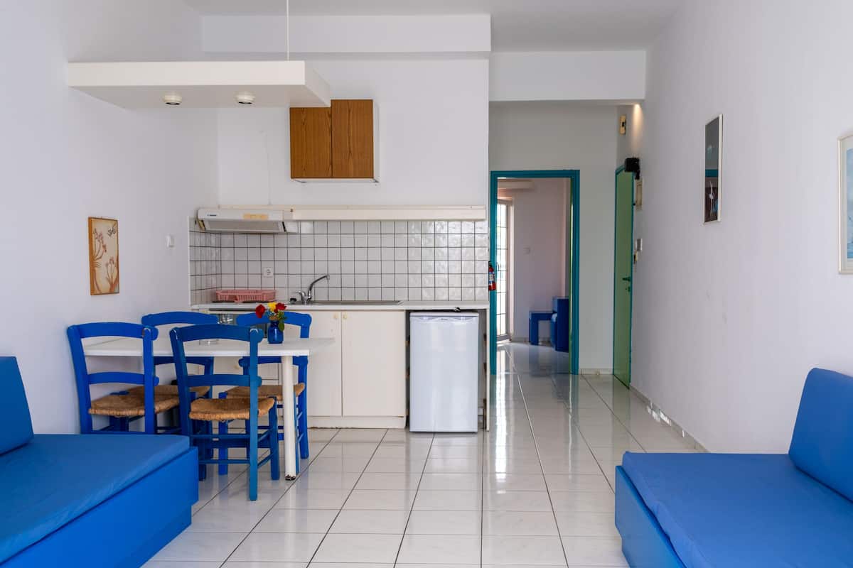 A modern kitchen with white cabinets, a built-in oven, and a sink. There are blue chairs and a dining table with a vase of flowers. A small refrigerator is visible, and the floor is tiled.