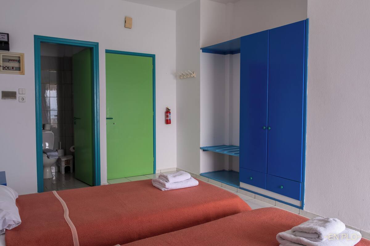A hotel room with two beds, a green door, a blue wardrobe, and a bathroom visible through an open door.